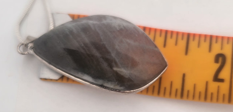 Metallic pendant with a dark stone inlay on a white background with a measuring tape for scale.