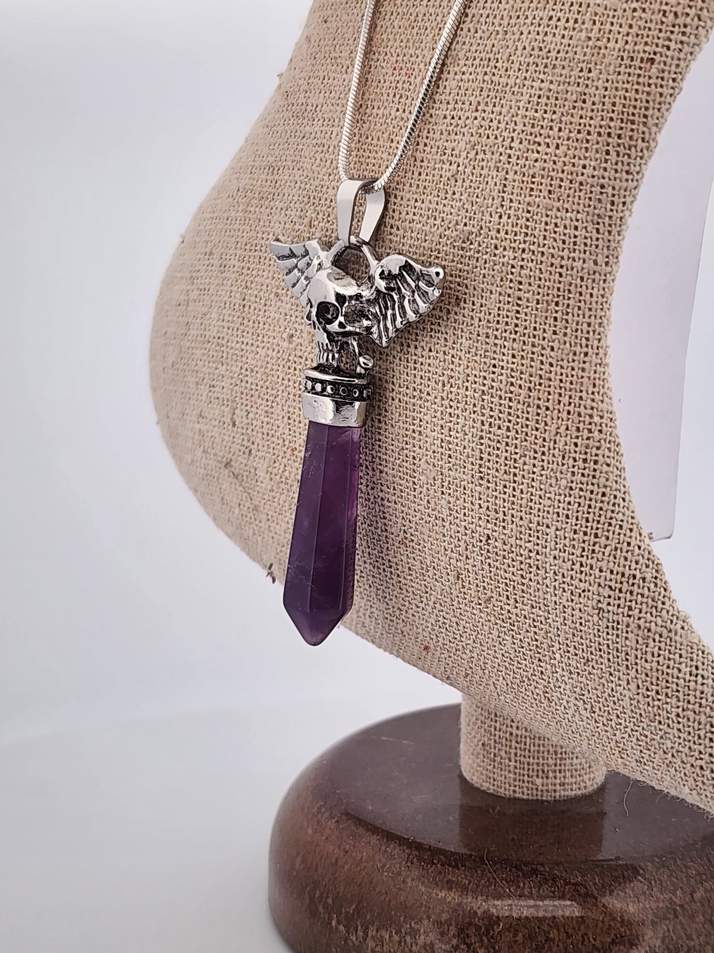 Necklace with a silver pendant and purple crystal on a beige stand.