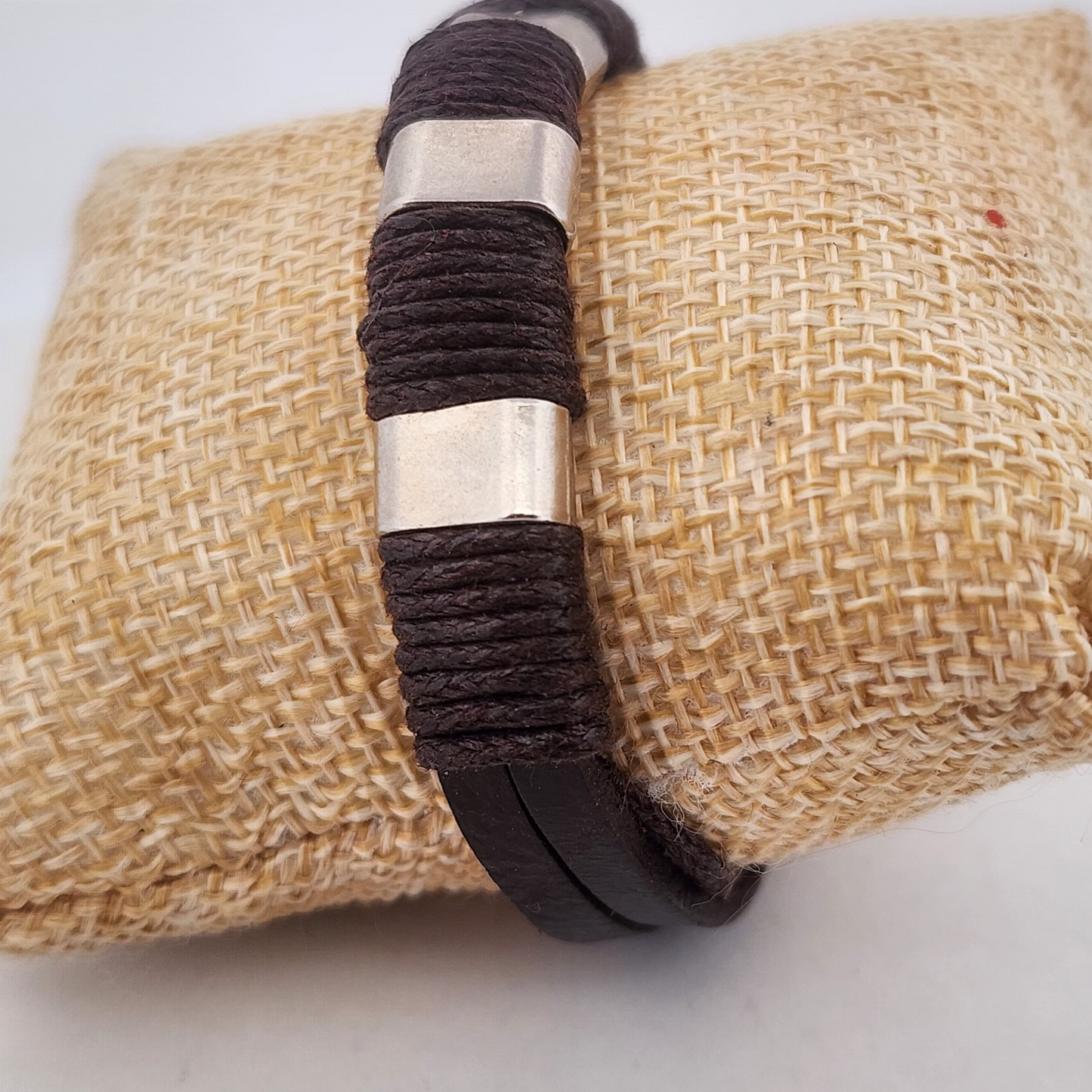 Bracelet with brown woven band and silver cylindrical center on a textured beige surface