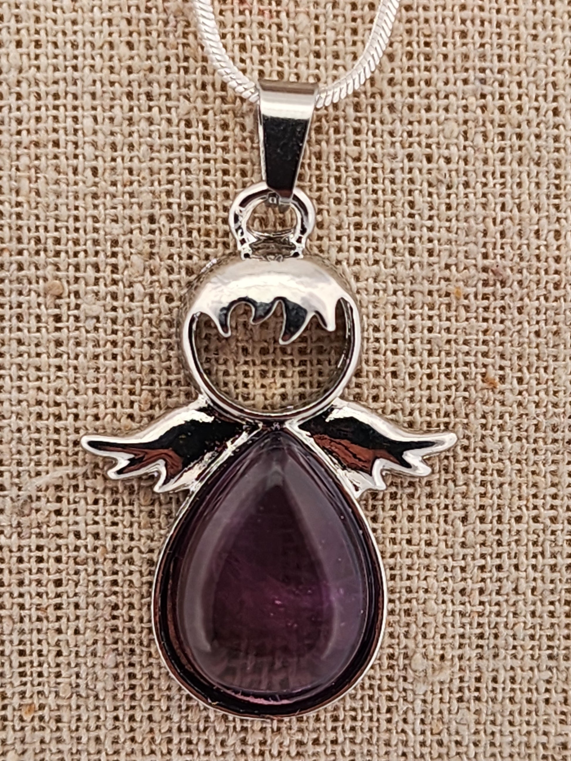 Silver angel pendant with a purple teardrop-shaped gemstone on a beige fabric background