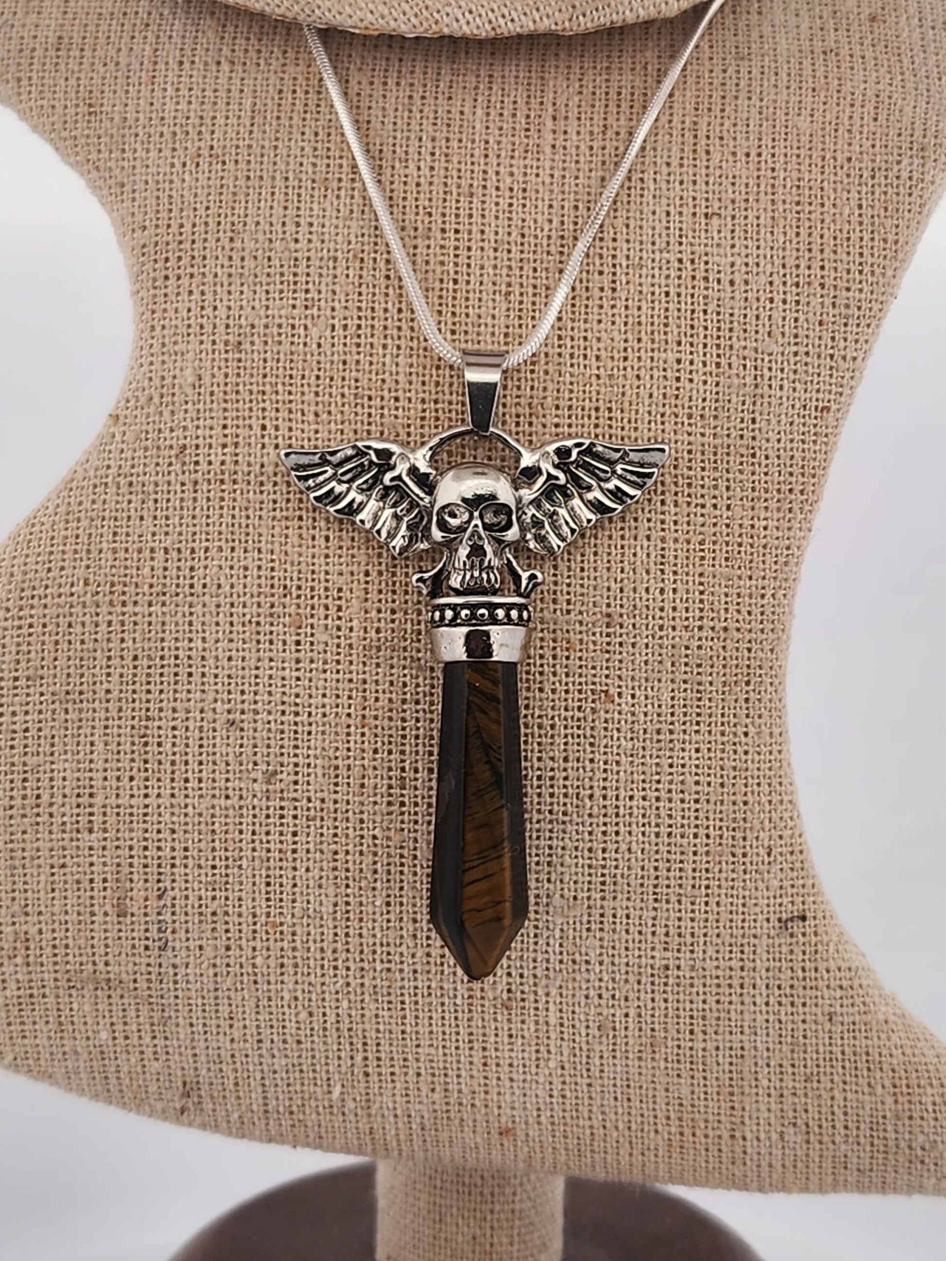 Skull pendant with wings and a brown gemstone on a beige display stand.