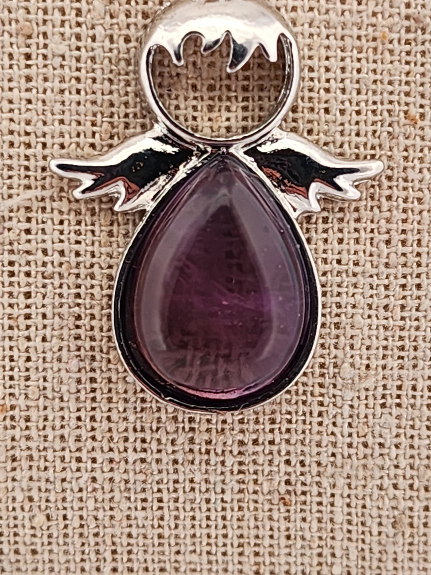 Purple teardrop pendant with silver wings on a beige fabric background