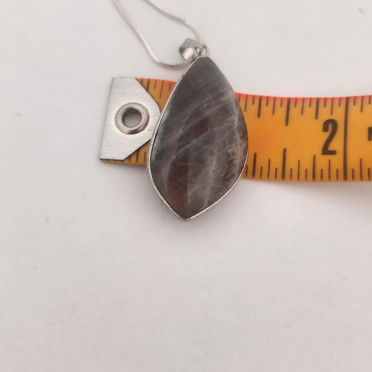 Teardrop-shaped pendant on a chain with a ruler for scale on a white background
