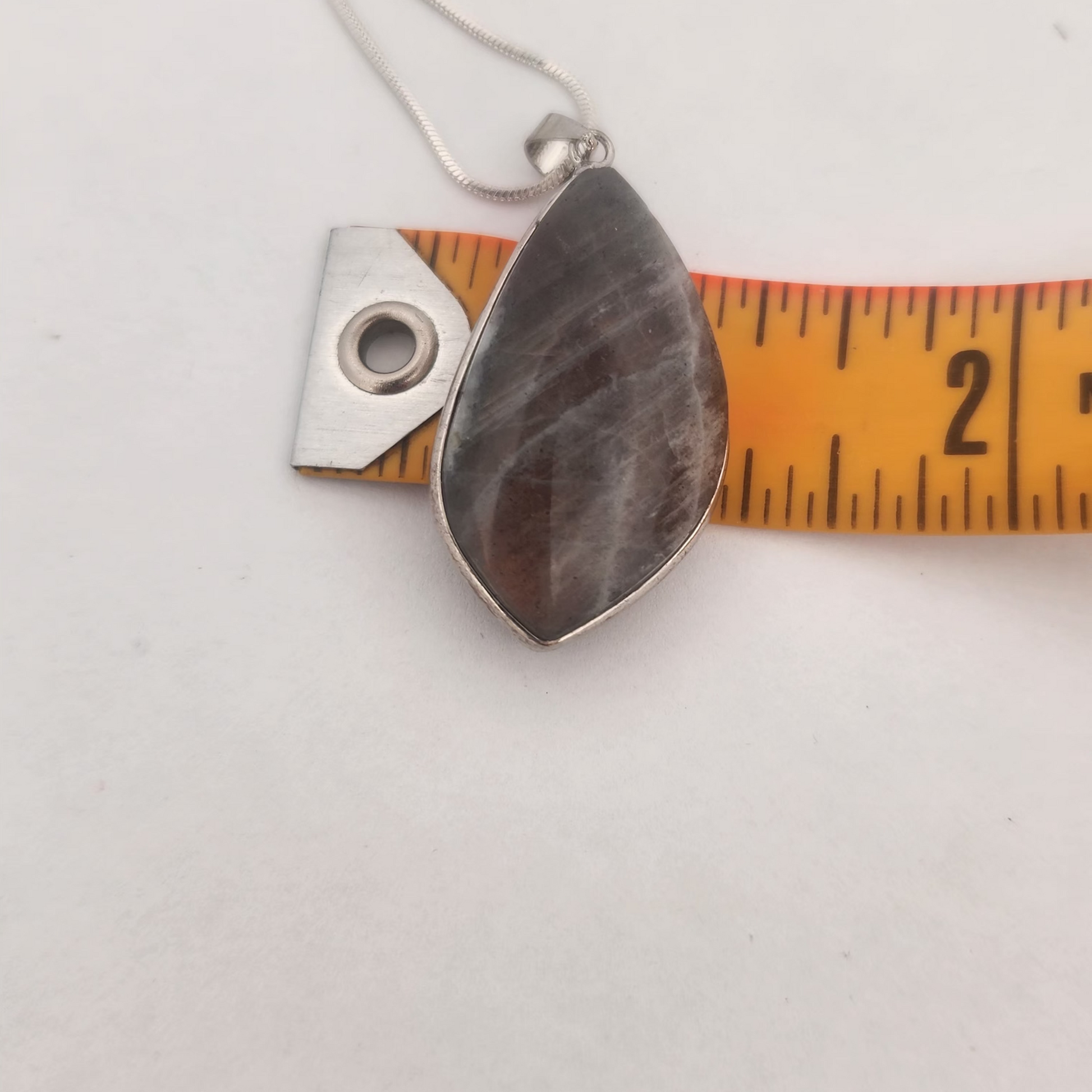 Teardrop-shaped pendant on a chain with a ruler for scale on a white background