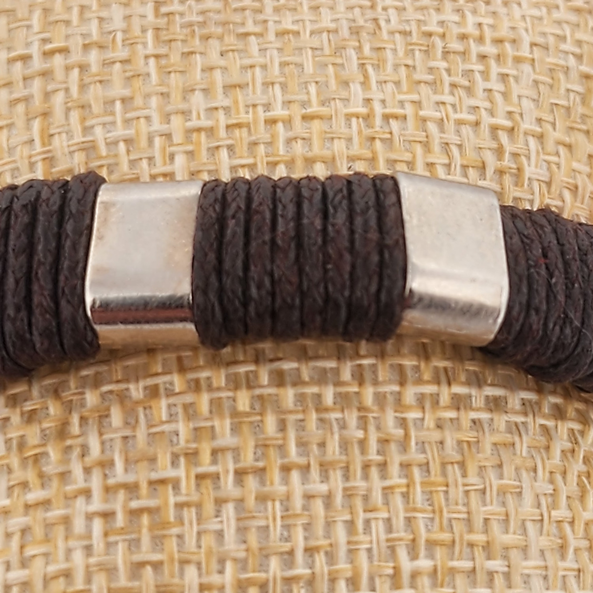 Close-up of a brown braided cord with a silver cylindrical metal piece on a textured beige background.